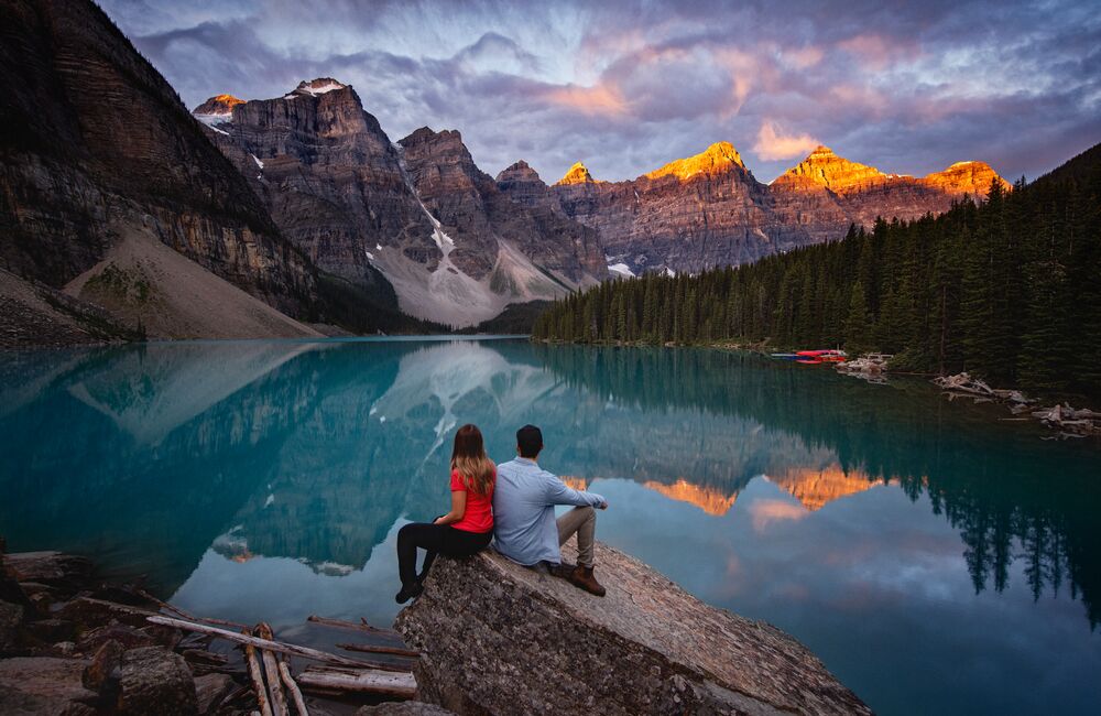 Two people sit on a rock by the water at Moraine Lake at sunrise in the summer. Alpen glow hits the tips of the mountain peaks and reflects in the water.